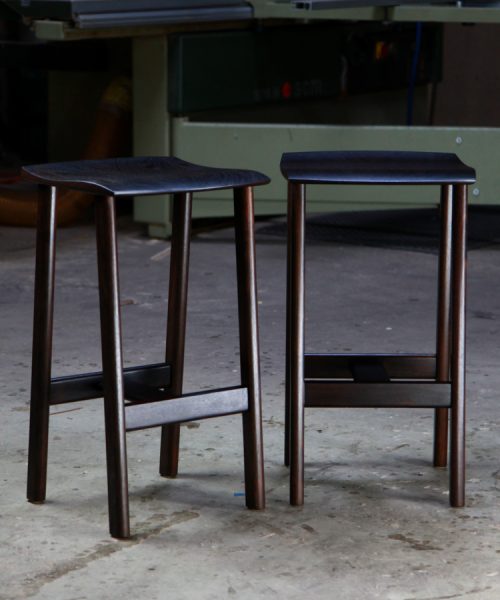 A moody photograph of two black timber stools in front of saw in a workshop.