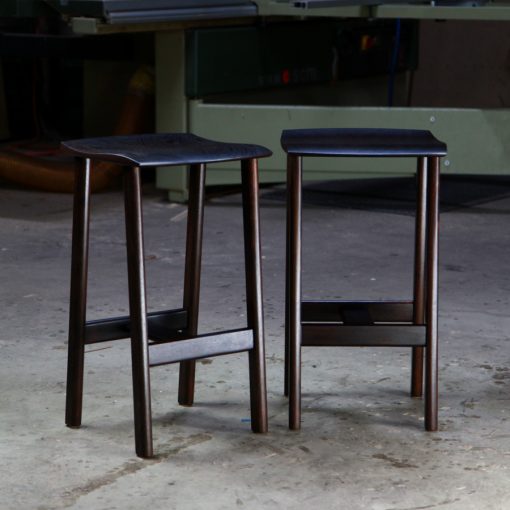 A moody photograph of two black timber stools in front of saw in a workshop.