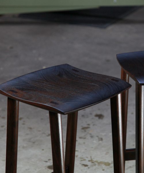 A moody photograph of two black timber stools in front of saw in a workshop.