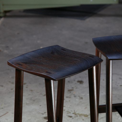 A moody photograph of two black timber stools in front of saw in a workshop.