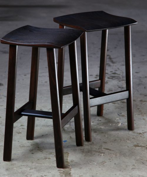 A moody photograph of two black timber stools in front of saw in a workshop.