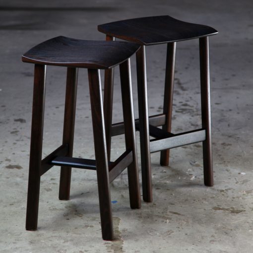 A moody photograph of two black timber stools in front of saw in a workshop.