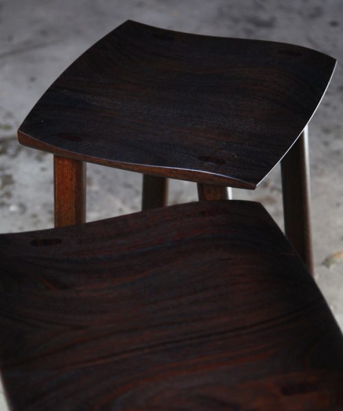 A moody photograph of two black timber stools in front of saw in a workshop.