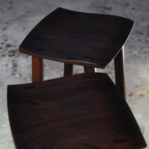 A moody photograph of two black timber stools in front of saw in a workshop.