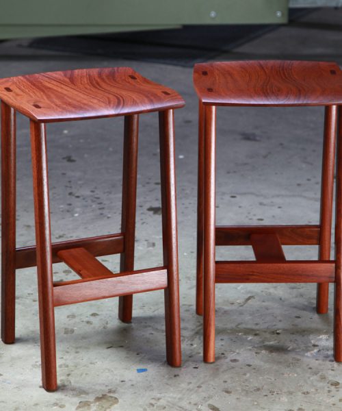 Two perfectly matched jarrah stools sit in a woodworking workshop.