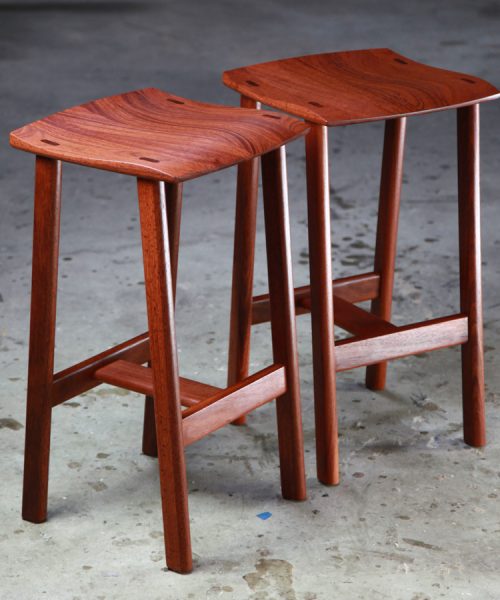 Two perfectly matched jarrah stools sit in a woodworking workshop.