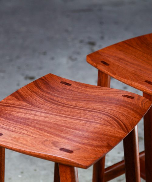 Two perfectly matched jarrah stools sit in a woodworking workshop.