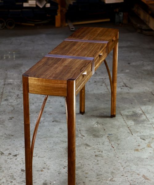 A beautifully composed hall table with figured blackwood standing in front of a timber rack in a woodworking workshop.