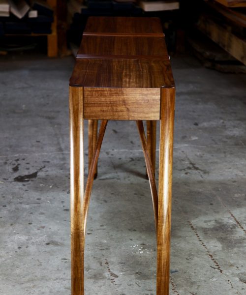 A beautifully composed hall table with figured blackwood standing in front of a timber rack in a woodworking workshop.