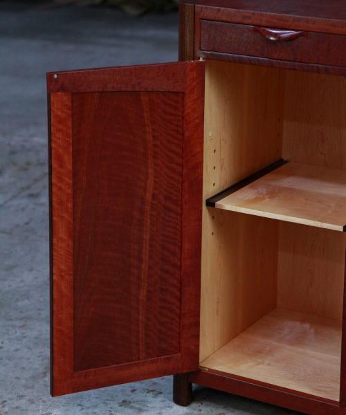 A beautiful red gum cabinet sits in front of a timber rack in a woodworking workshop.