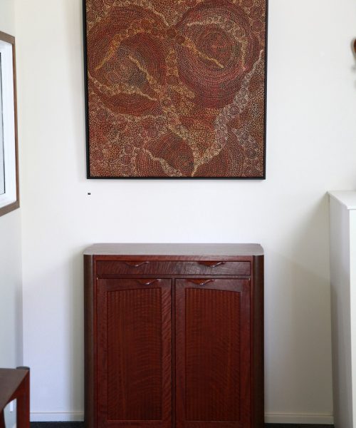 A cabinet in red gum underneath a painting by an Australian indigenous artist in a gallery.