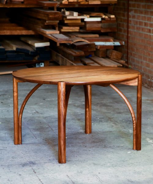 A beautifully composed round dining table in lightly figured blackwood standing in front of a timber rack in a woodworking workshop.