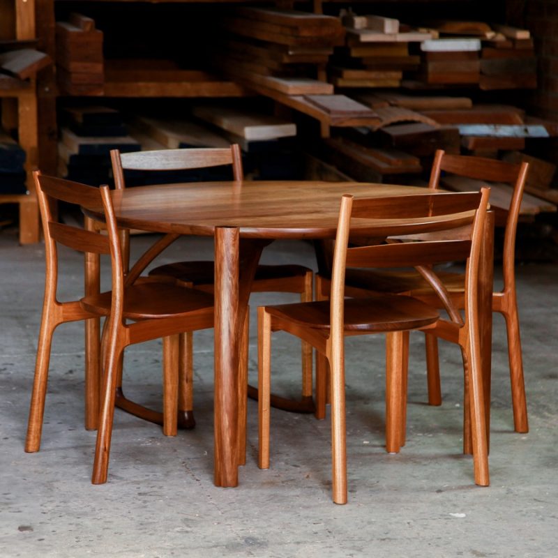 A beautifully composed round dining table with four chairs in lightly figured blackwood standing in front of a timber rack in a woodworking workshop.