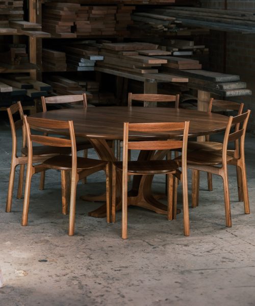 A round dining table with eight chairs in a woodworking workshop near a timber rack.