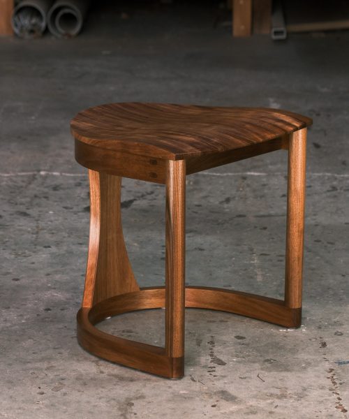 A low blackwood stools sit in a woodworking workshop close to a timber rack.
